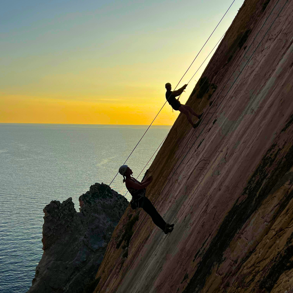 Two men abseiling down a rock face at sunset with yellow and blue sky tones.