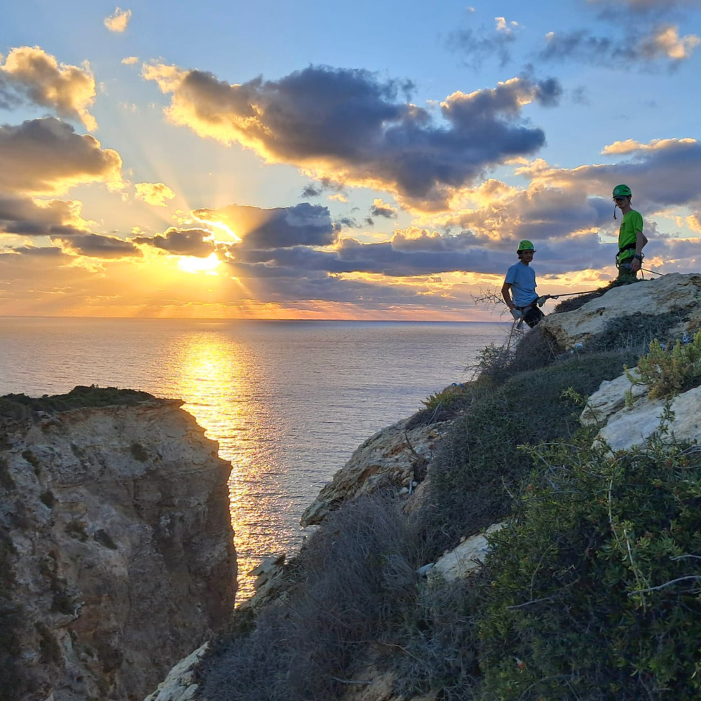Guide helping a client abseil down a rock face during sunset in Malta, both secured with ropes and safety gear.