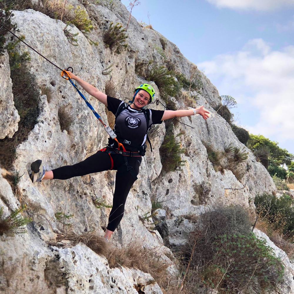 Woman posing on the Via Ferrata in Malta, clipped to the safety cable on the rock face.