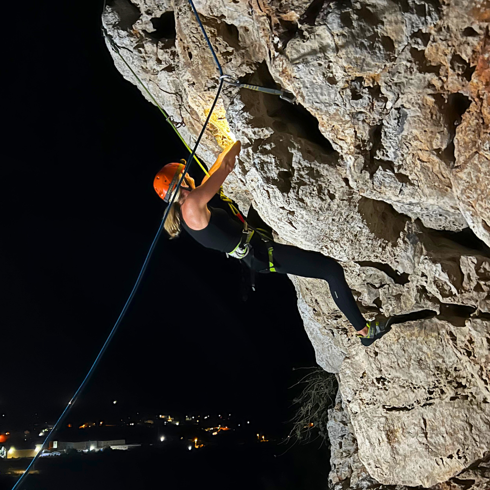 Woman climbing a rock face in Malta in the dark, wearing a harness and headlamp.